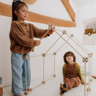 Des enfants jouent dans une cabane intérieure construite à l'aide du kit 1001 Cabanes Libellule de 43 pièces et de tissus ménagers, une construction en bois de hêtre à clipser qui stimule le jeu imaginatif et l'aventure.