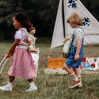 Due bambini che camminano all'aperto con i marsupi trapuntati Olli Ella sulla schiena, il marsupio verde salvia è chiaramente visibile mentre i bambini giocano insieme incoraggiando la cura condivisa e il gioco sociale.