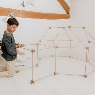 Des enfants à l'intérieur d'un grand fort construit à l'intérieur à partir du kit de 72 pièces « 1001 Cabanes Écureuil » et de draps ; cette cabane en bois de hêtre à assembler par emboîtement favorise le travail d'équipe et le jeu collaboratif