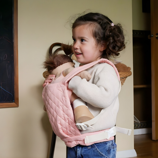 Child wearing Olli Ella quilted doll carrier in rose pink with doll secured in front, adjustable buckle straps and padded quilted cotton visible, hands-free baby carrier for nurturing role play