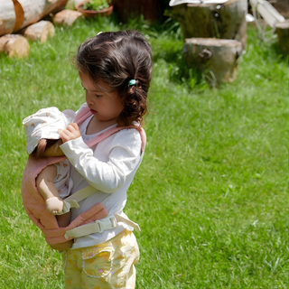 Child carrying doll in Olli Ella quilted carrier in rose pink while walking outdoors in garden, hands-free cotton baby carrier encouraging caring play and outdoor adventures with dolls