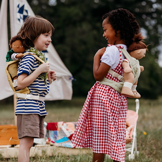 Two children outdoors each wearing Olli Ella quilted doll carriers with dolls on their backs, sage green and mustard carriers shown from behind during imaginative outdoor adventure play
