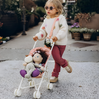 Child pushing Olli Ella Sollie Stroller in rose pink along pavement with doll seated inside, lightweight folding doll pushchair with working wheels for outdoor caregiving adventures and walks